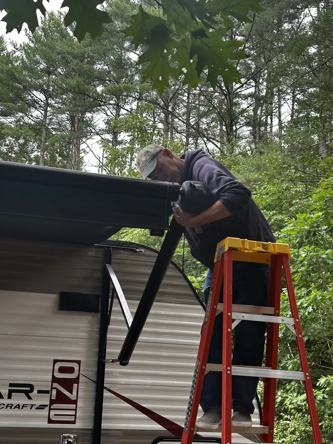 RV awning technician on a stepladder fitting a new patio awning to the side of a travel trailer at a wooded campsite
