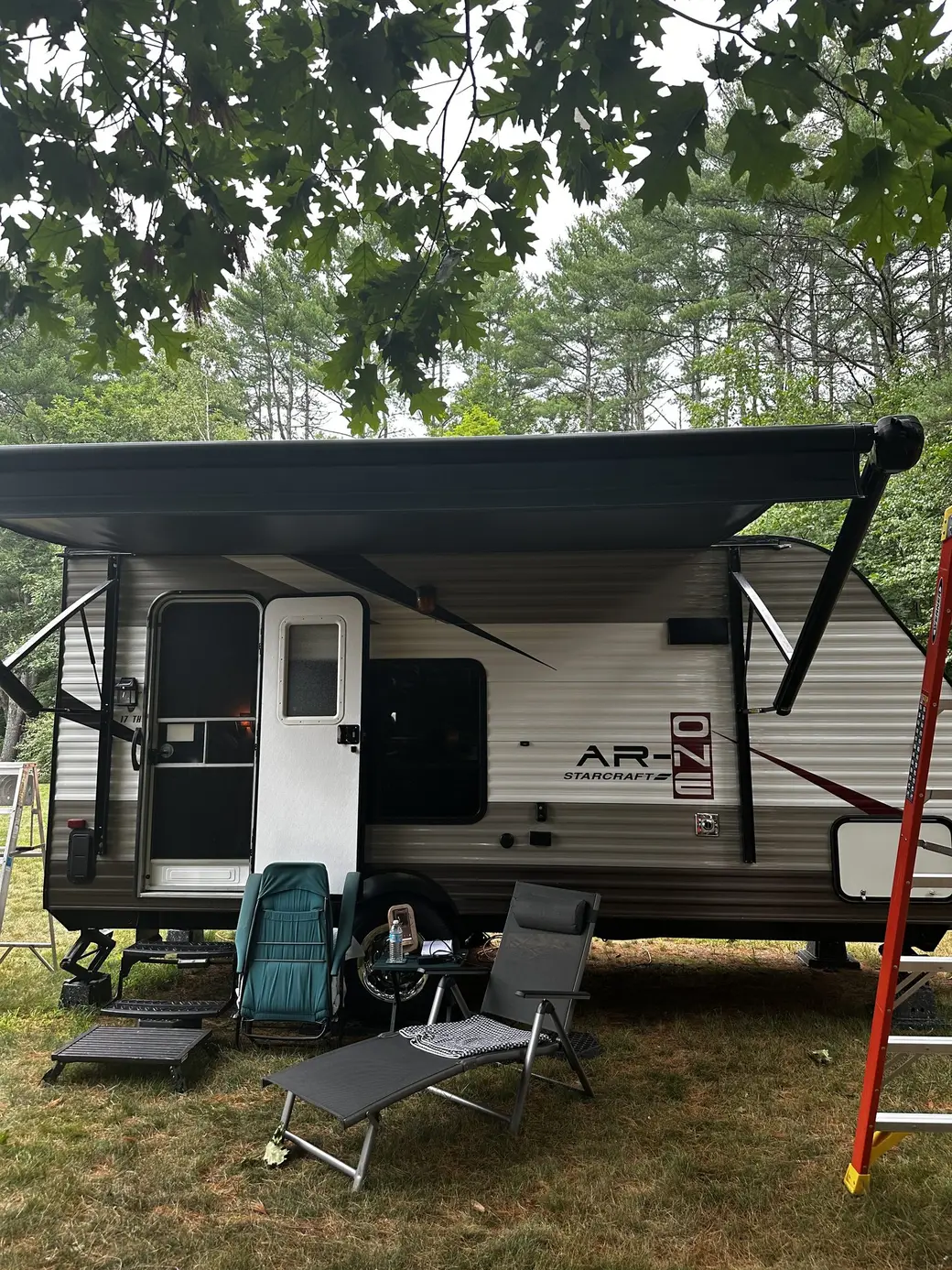Newly installed RV patio awning extended over a travel trailer at a wooded campsite, with camp chairs set up in the shade underneath